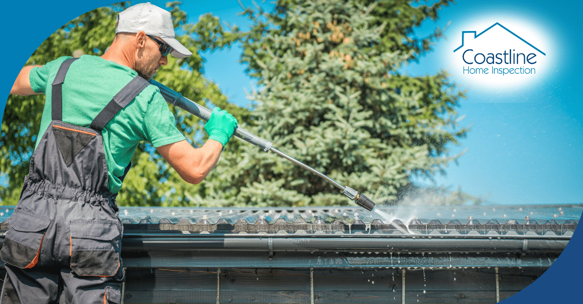 A professional in green attire and gloves uses a pressure washer to clean the roof and gutter system of a home under a clear blue sky.